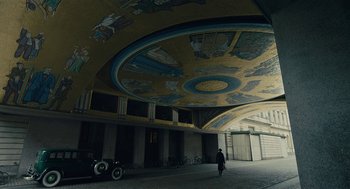 Movie still from “The Danish Girl” (2015), directed by Tom Hooper – A man walking in a building with a car parked in front of it; Extreme Wide shot, Low angle