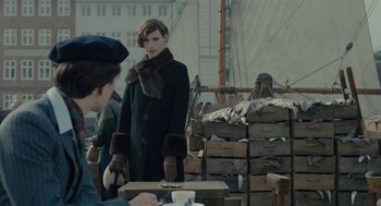 Movie still from “The Danish Girl” (2015), directed by Tom Hooper – A woman in a black coat standing in front of a table; Medium shot, Over the shoulder angle