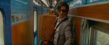 Movie still from “The Darjeeling Limited” (2007), directed by Wes Anderson – A man in a suit and sunglasses holding a suitcase; Medium shot, Low angle