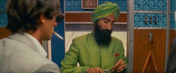 Movie still from “The Darjeeling Limited” (2007), directed by Wes Anderson – A man in a green suit and a yellow turban smoking a cigarette and looking at his cell phone; Medium shot, Over the shoulder angle