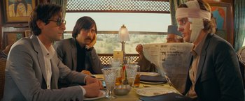Movie still from “The Darjeeling Limited” (2007), directed by Wes Anderson – A group of people sitting at a table with a newspaper; Medium shot, Over the shoulder angle