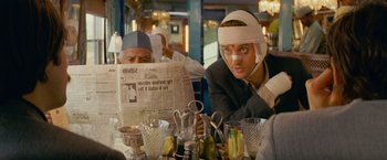 Movie still from “The Darjeeling Limited” (2007), directed by Wes Anderson – A man with a head injury sitting at a table with other people; Medium shot, Over the shoulder angle