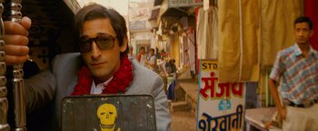 Movie still from “The Darjeeling Limited” (2007), directed by Wes Anderson – A man in a suit and a red scarf is holding a suitcase; Close Up shot, Over the shoulder angle