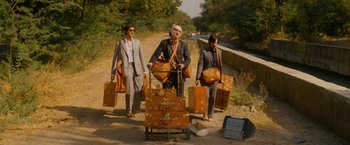 Movie still from “The Darjeeling Limited” (2007), directed by Wes Anderson – A group of people standing next to suitcases on the side of a road; Wide shot, Low angle
