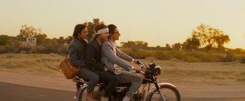 Movie still from “The Darjeeling Limited” (2007), directed by Wes Anderson – A group of men riding on the back of a motorcycle down a street; Wide shot, Low angle