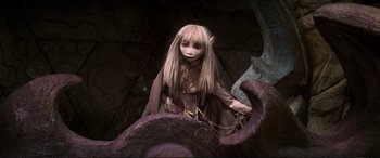 Movie still from “The Dark Crystal” (1982), directed by Frank Oz – A young girl dressed as an elf sitting on top of a tree trunk; Medium shot, High angle