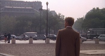 Movie still from “The Day of the Jackal” (1973), directed by Fred Zinnemann – A man in a brown jacket standing in a parking lot next to parked cars; Wide shot, Over the shoulder angle