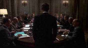 Movie still from “The Day of the Jackal” (1973), directed by Fred Zinnemann – A man standing in front of a group of people at a table; Wide shot, Over the shoulder angle