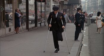 Movie still from “The Day of the Jackal” (1973), directed by Fred Zinnemann – An older man in a suit and a hat is on crutches; Wide shot, High angle