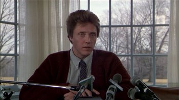 Movie still from “The Dead Zone” (1983), directed by David Cronenberg – A man sitting at a table with microphones in front of him; Close Up shot, Low angle