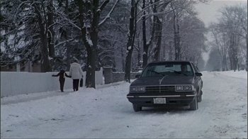 Movie still from “The Dead Zone” (1983), directed by David Cronenberg – Two people walk down a snow covered road past a car; Wide shot, High angle