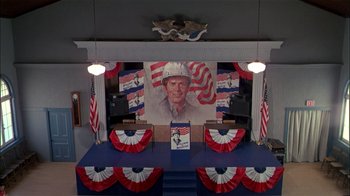 Movie still from “The Dead Zone” (1983), directed by David Cronenberg – A large portrait of a man in a hat on a stage; Wide shot, High angle