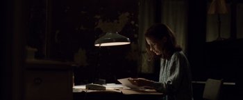 Movie still from “The Debt” (2010), directed by John Madden – A woman sitting at a desk looking at a piece of paper in front of a lamp; Medium shot, Over the shoulder angle