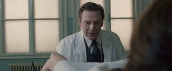 Movie still from “The Debt” (2010), directed by John Madden – A man in a white lab coat sitting in front of a table; Close Up shot, Over the shoulder angle