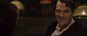 Movie still from “The Debt” (2010), directed by John Madden – A person wearing a suit and smiling; Close Up shot, Over the shoulder angle