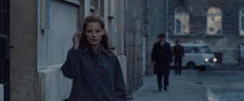Movie still from “The Debt” (2010), directed by John Madden – A woman walking down a street with a man in the background; Medium shot, Over the shoulder angle