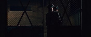 Movie still from “The Debt” (2010), directed by John Madden – A couple of men standing next to each other in front of a chain link fence; Wide shot, Low angle