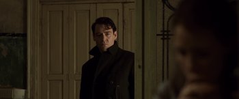 Movie still from “The Debt” (2010), directed by John Madden – A man in a black coat standing in front of a door; Medium shot, Low angle