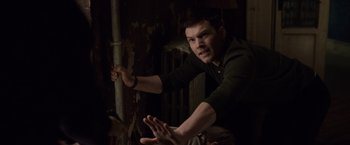Movie still from “The Debt” (2010), directed by John Madden – A man leaning against a pole in a dark room; Medium shot, Over the shoulder angle