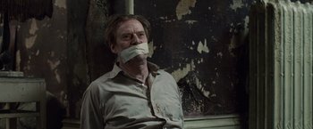 Movie still from “The Debt” (2010), directed by John Madden – A man with his mouth taped shut is sitting in front of a wall; Close Up shot, High angle