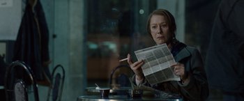 Movie still from “The Debt” (2010), directed by John Madden – A woman sitting at a table holding a cigarette and a newspaper; Medium shot, Over the shoulder angle