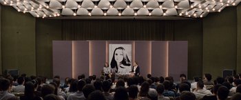 Movie still from “The Debt” (2010), directed by John Madden – Two people sitting in front of an audience in front of a picture of a woman; Wide shot, Over the shoulder angle