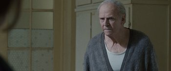 Movie still from “The Debt” (2010), directed by John Madden – An older man in a white shirt and a gray sweater; Medium shot, Low angle