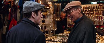 Movie still from “The Devil's Advocate” (1997), directed by Taylor Hackford – Two men are talking in front of a display of gold jewelry; Medium shot, Over the shoulder angle