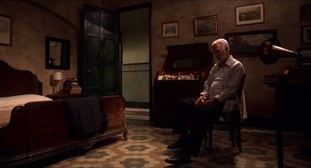 Movie still from “The Devil's Backbone” (2001), directed by Guillermo del Toro – An older man sitting on a chair in a living room; Wide shot, High angle