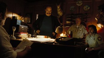 Movie still from “The Devil's Rejects” (2005), directed by Rob Zombie – A man standing in front of a table with a lamp on top of it; Medium shot, Low angle