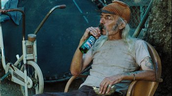 Movie still from “The Devil's Rejects” (2005), directed by Rob Zombie – An older man sitting in a chair drinking a beer; Medium shot, High angle