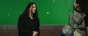Movie still from “The Disaster Artist” (2017), directed by James Franco – A man with long black hair standing in front of a green wall; Medium shot, Over the shoulder angle