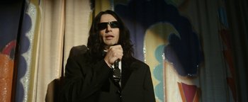 Movie still from “The Disaster Artist” (2017), directed by James Franco – A man with long black hair wearing sunglasses and holding a microphone; Medium shot, Low angle