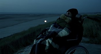 Movie still from “The Diving Bell and the Butterfly” (2007), directed by Julian Schnabel – A man sitting in a wheelchair on the beach at night; Medium shot, Over the shoulder angle
