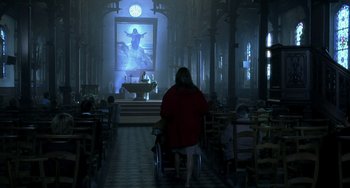 Movie still from “The Diving Bell and the Butterfly” (2007), directed by Julian Schnabel – A woman in a wheelchair in front of an altar with an image of jesus in the background; Wide shot, Low angle