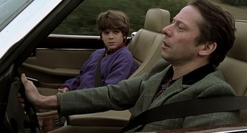 Movie still from “The Diving Bell and the Butterfly” (2007), directed by Julian Schnabel – A man driving a car while a boy sits in the driver's seat; Medium shot, Over the shoulder angle