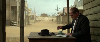Movie still from “The Dressmaker” (2015), directed by Jocelyn Moorhouse – A man is drinking coffee in front of an old western town; Wide shot, Over the shoulder angle