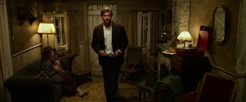 Movie still from “The Dressmaker” (2015), directed by Jocelyn Moorhouse – A man standing in a living room holding a book; Wide shot, Low angle