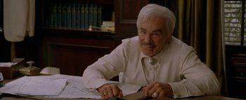 Movie still from “The Dukes of Hazzard” (2005), directed by Jay Chandrasekhar – An older man sitting at a table cutting paper with a knife; Close Up shot, High angle