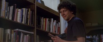 Movie still from “The End of the Tour” (2015), directed by James Ponsoldt – A man holding a cell phone in front of a bookshelf; Medium shot, Over the shoulder angle
