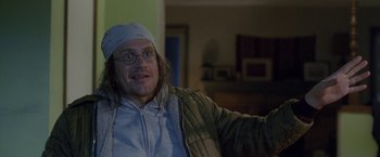 Movie still from “The End of the Tour” (2015), directed by James Ponsoldt – A man wearing glasses and a hat sitting in a living room; Close Up shot, Over the shoulder angle