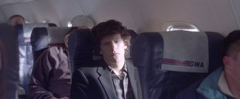 Movie still from “The End of the Tour” (2015), directed by James Ponsoldt – A man sitting on a seat on a plane looking at the camera; Medium shot, Over the shoulder angle