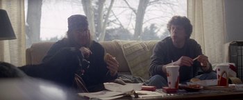 Movie still from “The End of the Tour” (2015), directed by James Ponsoldt – Two people sitting on a couch eating a hot dog; Medium shot, Over the shoulder angle