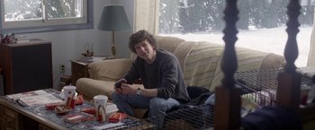 Movie still from “The End of the Tour” (2015), directed by James Ponsoldt – A man sitting on a couch holding a nintendo wii game controller; Medium shot, Over the shoulder angle