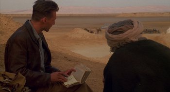 Movie still from “The English Patient” (1996), directed by Anthony Minghella – A man sitting on the side of a road while holding a book; Medium shot, Over the shoulder angle