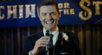 Movie still from “The Fabelmans” (2022), directed by Steven Spielberg – A man in a suit and tie holding a microphone and smiling; Close Up shot, Over the shoulder angle