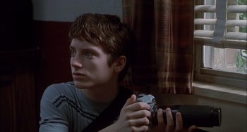 Movie still from “The Faculty” (1998), directed by Robert Rodriguez – A young man holding a camera while sitting in a room; Close Up shot, Low angle