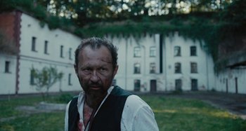 Movie still from “The Field Guide to Evil” (2018), directed by Can Evrenol – A man with a beard and a white shirt is standing in front of a white building; Medium shot, Low angle