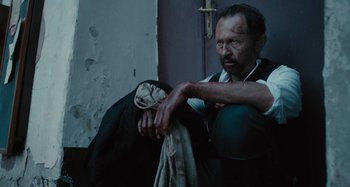Movie still from “The Field Guide to Evil” (2018), directed by Can Evrenol – A man sitting next to a door with a knife in his hand; Medium shot, High angle