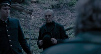 Movie still from “The Field Guide to Evil” (2018), directed by Can Evrenol – An older man wearing a black coat and a black hat; Medium shot, Over the shoulder angle